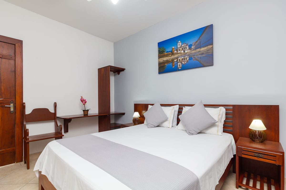 A hotel room with a bed, bedside tables, a chair, a wooden wardrobe, a painting on the wall, and a small table with a lamp.