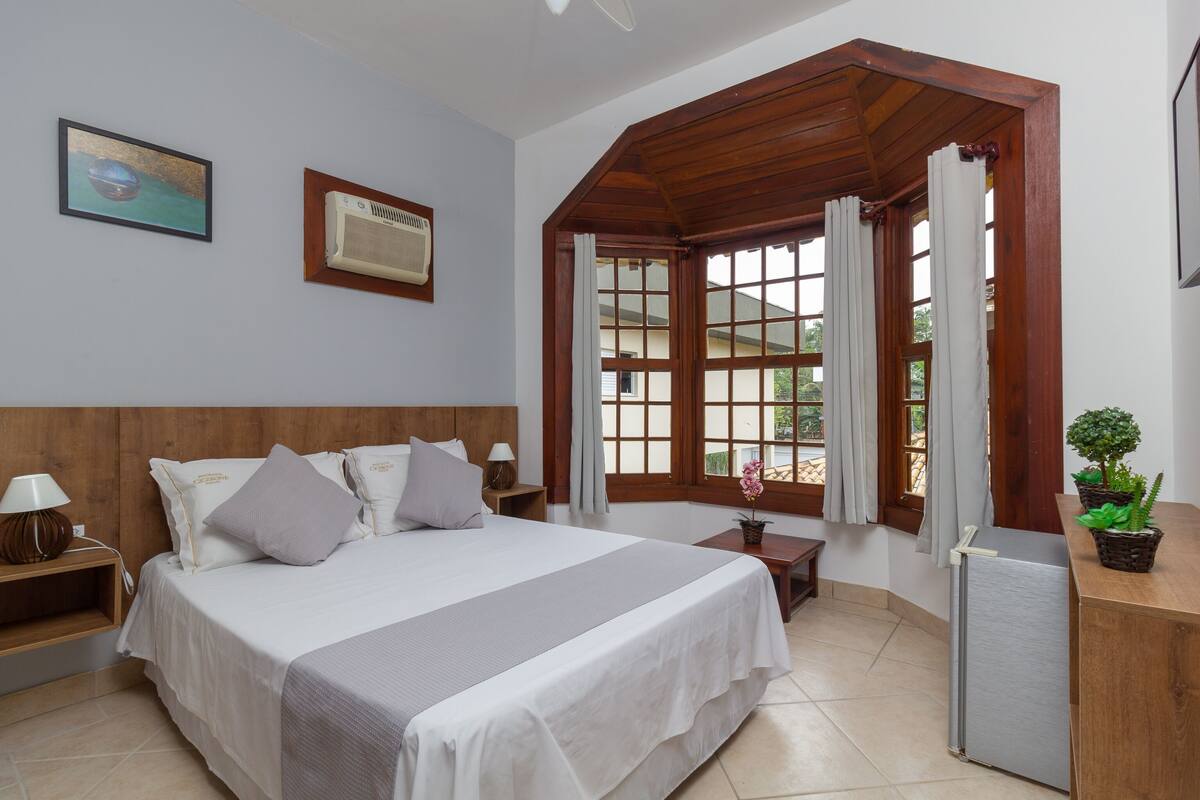 A bedroom with a bed, bedside tables, a desk, a refrigerator, a window with curtains, and a potted plant.
