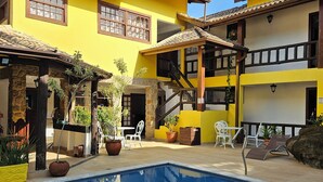 Zona de piscina con una piscina azul cristalina, edificios amarillos y mobiliario exterior.