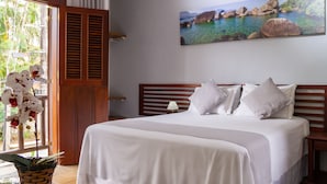 Un dormitorio con cama, almohadas, una mesita de noche, lámpara, ventana con persianas y un cuadro de un paisaje costero.