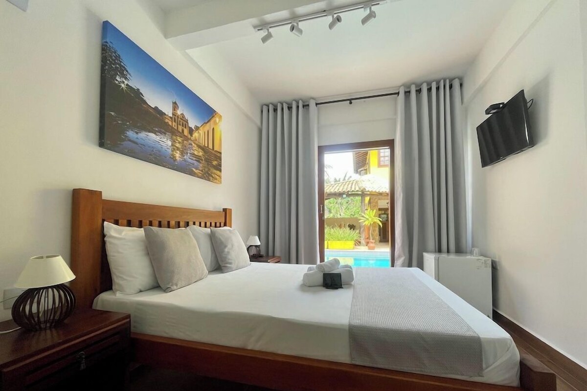 A hotel room with a bed, a nightstand, a lamp, a painting on the wall, a TV mounted on the wall, and a view of an outdoor area with a pool.