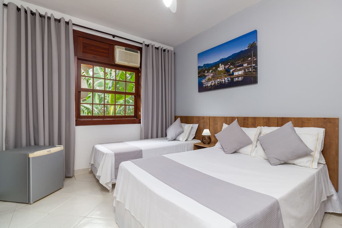 A hotel room with two beds, a window with curtains, a wall-mounted air conditioner, and a painting of a coastal town.