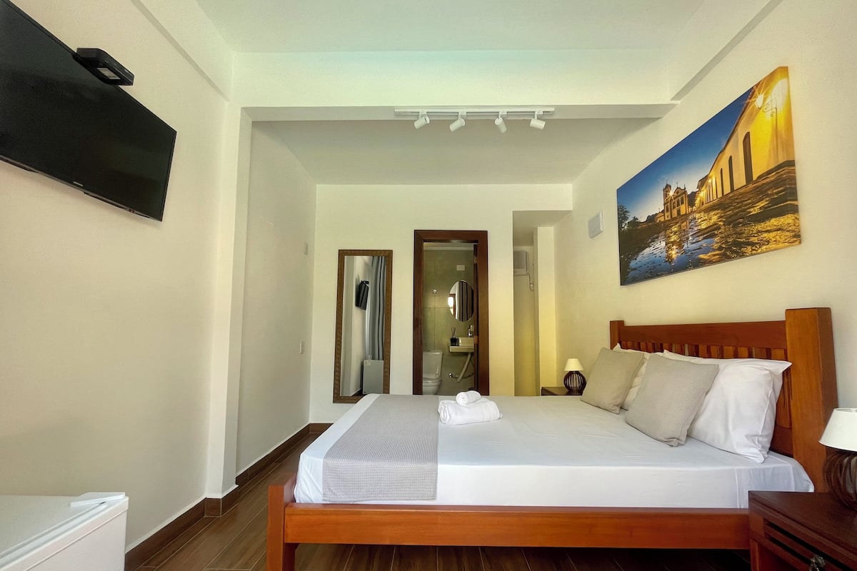 A hotel room with a bed, a TV, a painting, and a bathroom visible through an open door.