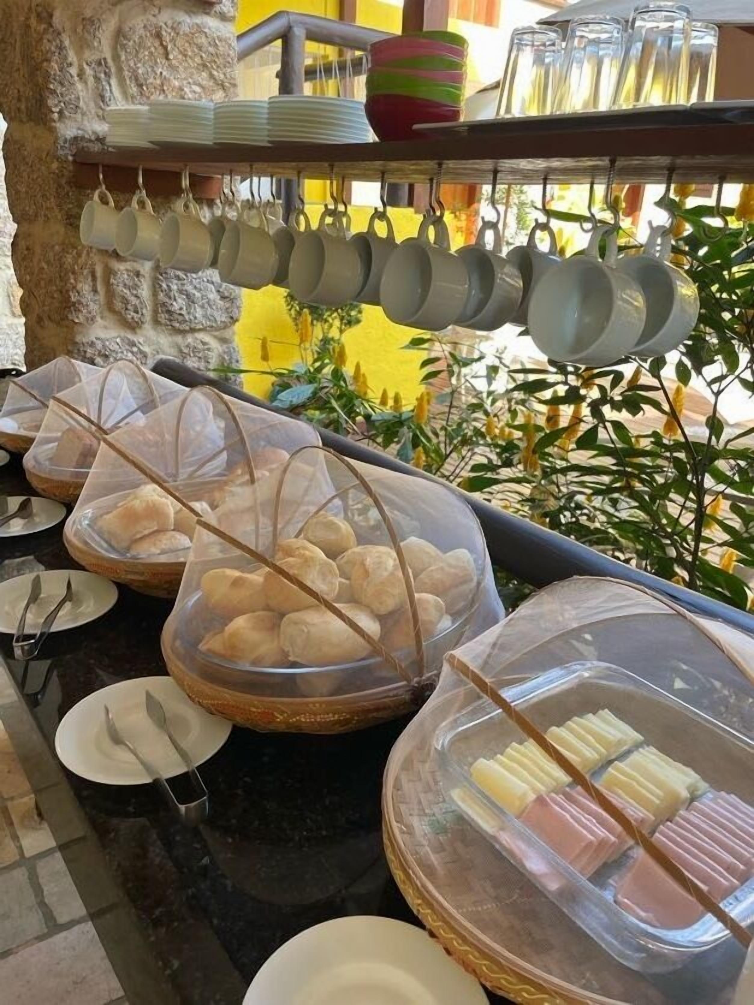 A buffet setup with clear plastic covers over food items, a variety of plates and cups, and a stone wall background.