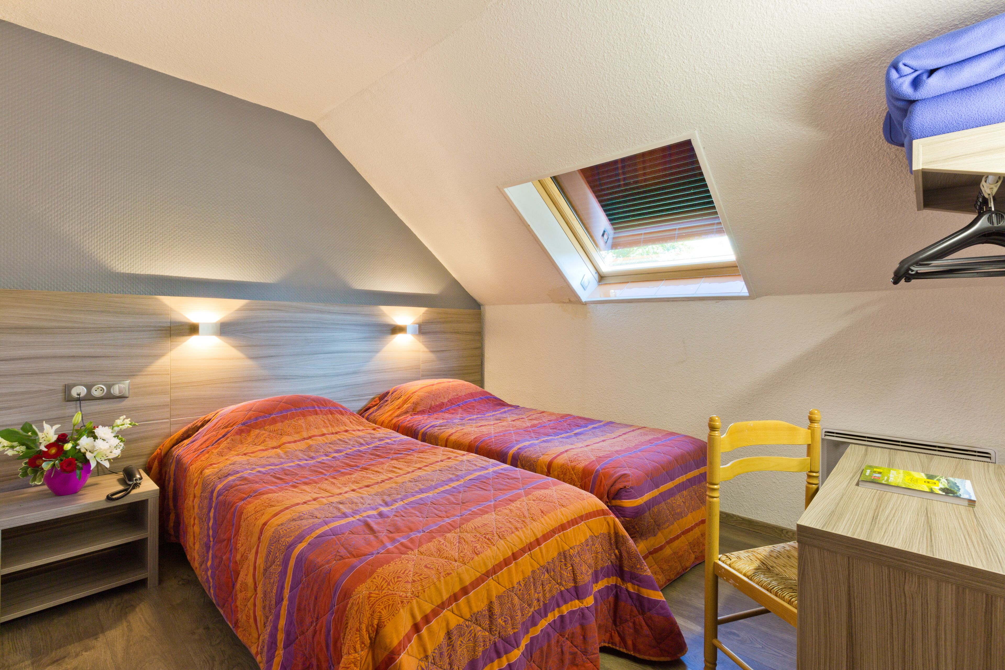 A small bedroom with two beds, a desk, and a chair. There is a skylight above the desk.