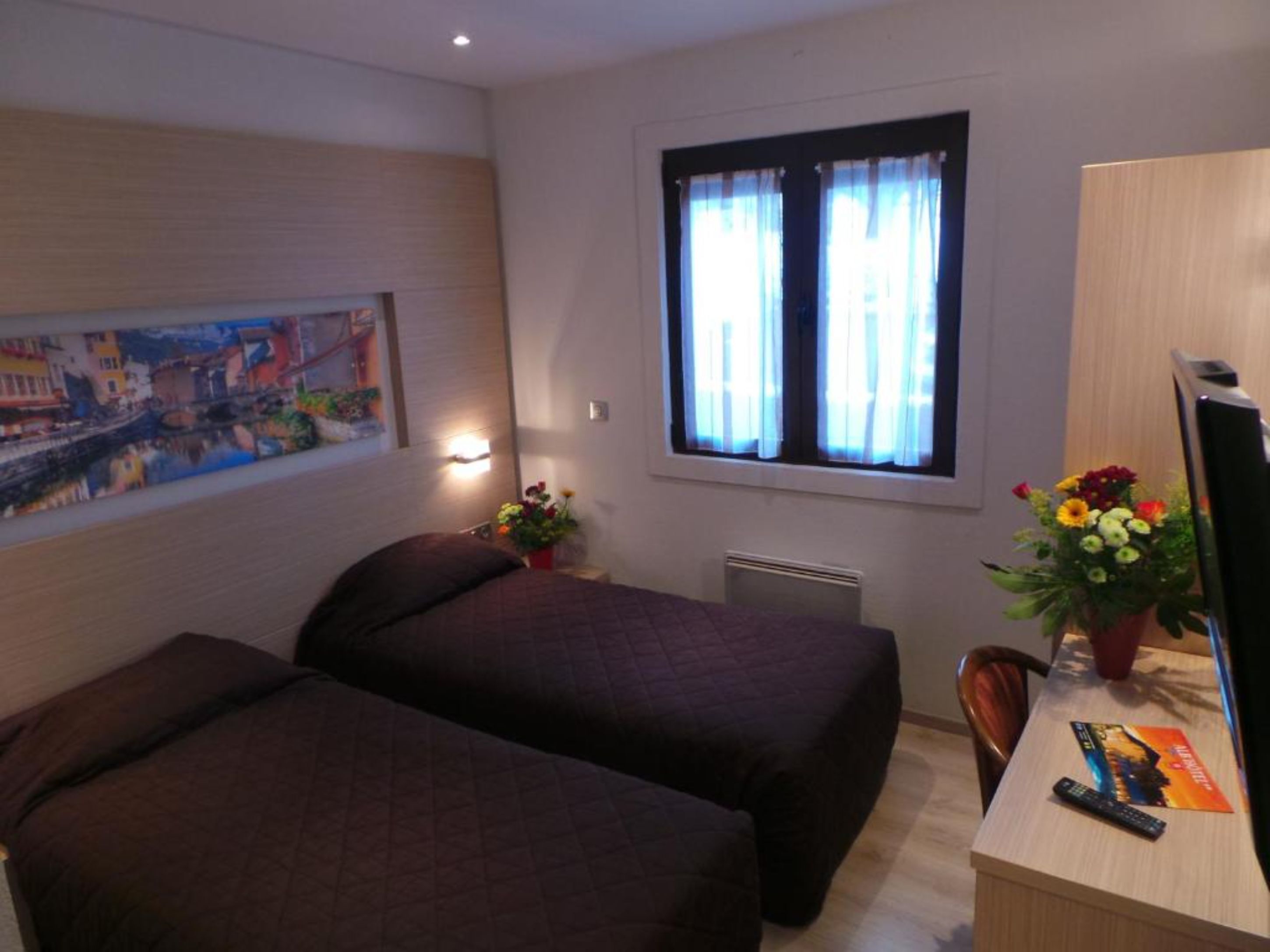 A hotel room with two beds, a desk with a computer and a remote control, a window with curtains, a painting on the wall, and a vase with flowers.