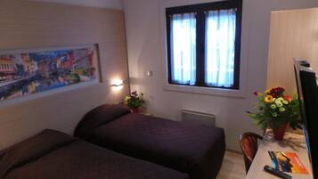 A hotel room with two beds, a desk with a computer and a remote control, a window with curtains, a painting on the wall, and a vase with flowers.