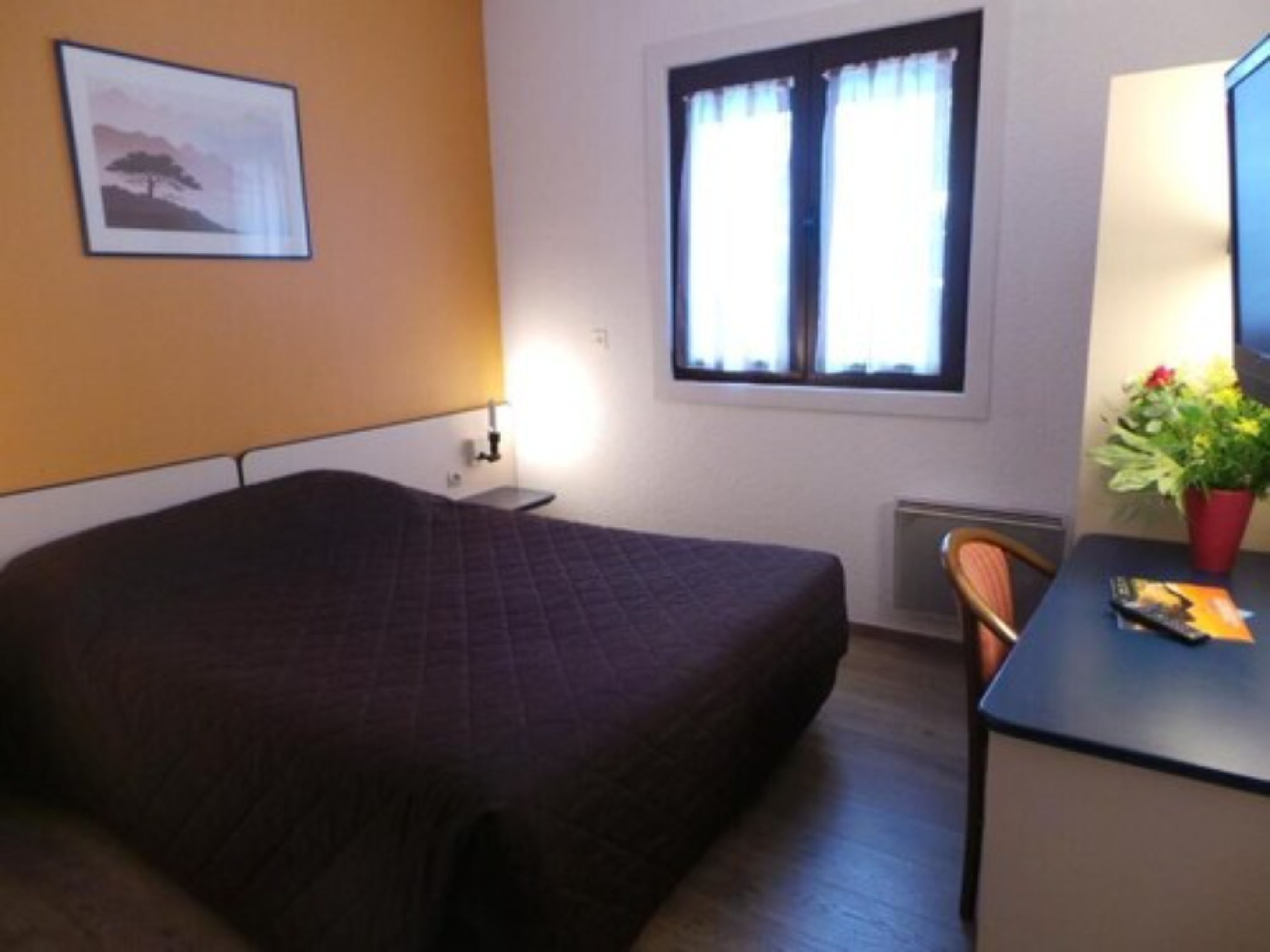 Une chambre d’hôtel avec un lit, un bureau, une chaise, une fenêtre avec des rideaux, un radiateur et un tableau encadré au mur.
