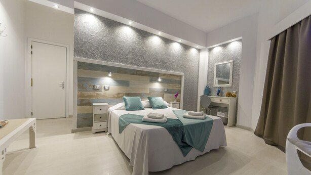A modern bedroom with a large bed, bedside tables, a vanity with a mirror, and a door leading to another room. - Hotel Sun Holidays (Puerto de la Cruz)
