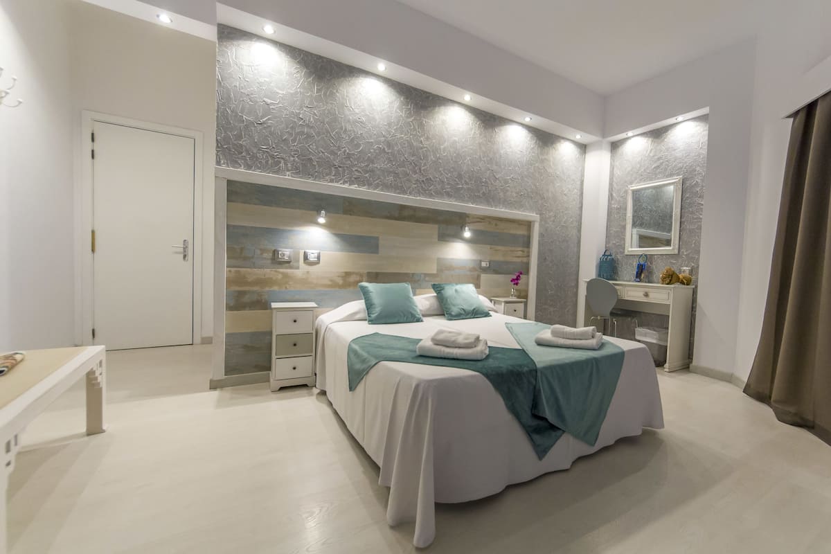 Quarto moderno com uma cama grande, mesas de cabeceira, toucador com espelho e uma porta que dá para outro cômodo.