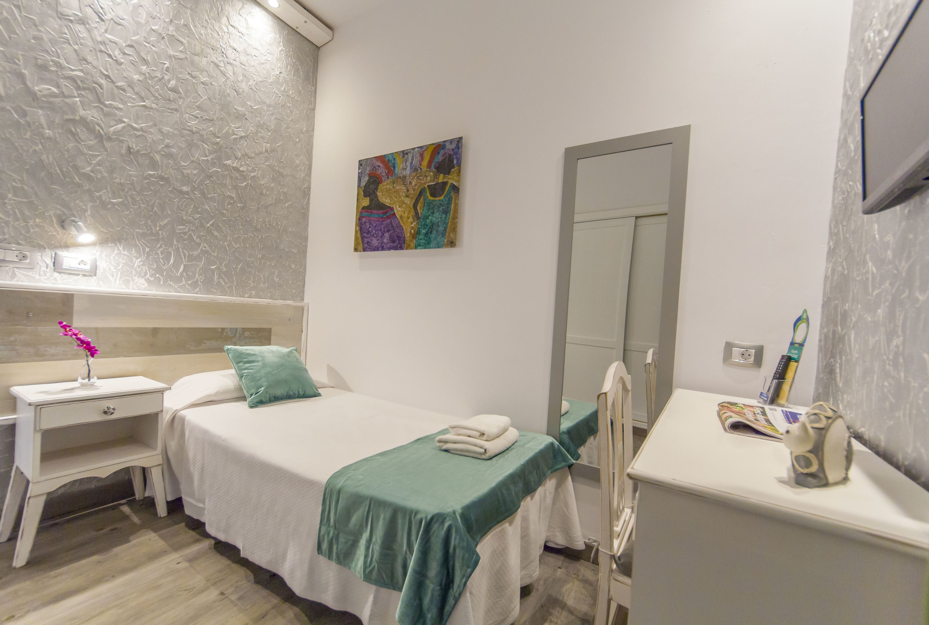 A hotel room with a bed, a nightstand, a mirror, a painting, and a small table with a toothbrush and a cup.
