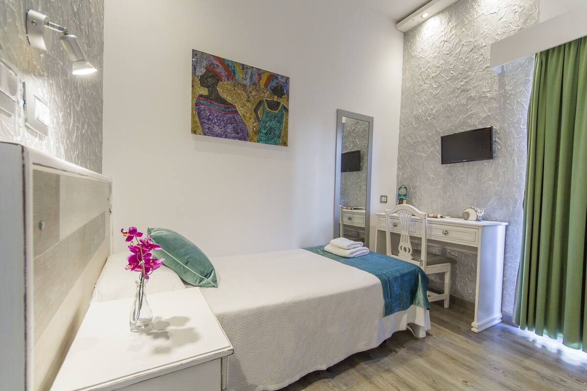 Quarto com cama, mesa de cabeceira com um vaso de flores, cômoda, espelho e televisão fixada na parede.