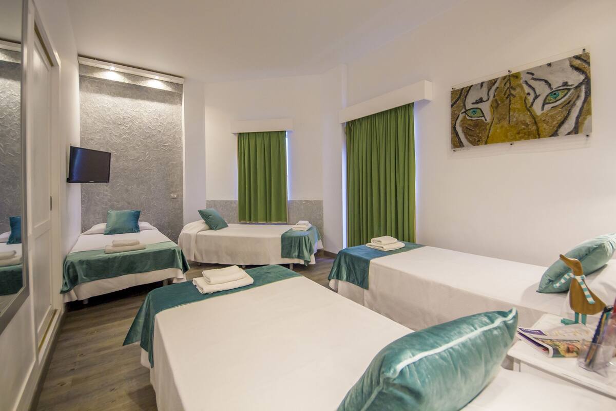 Quarto de hotel com duas camas de solteiro, televisão, um quadro e janela com cortinas verdes.