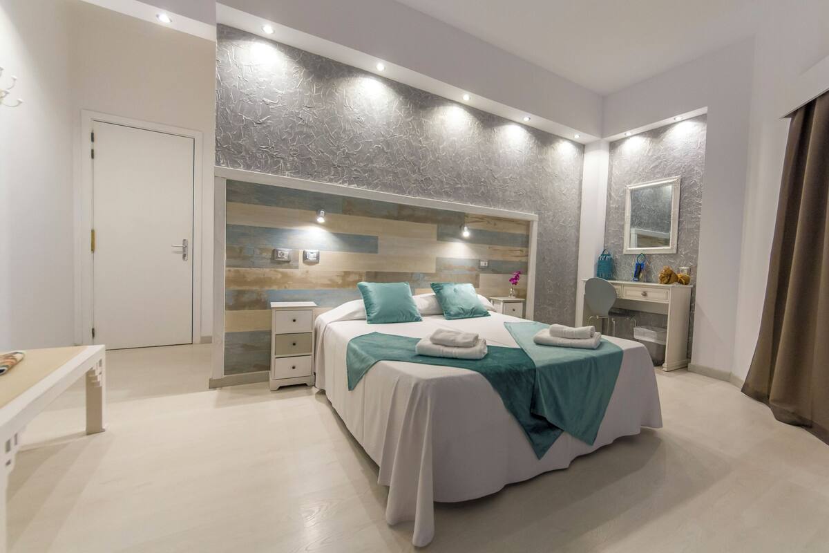 Quarto moderno com uma cama grande, mesas de cabeceira, toucador com espelho e uma porta que dá para outro cômodo.