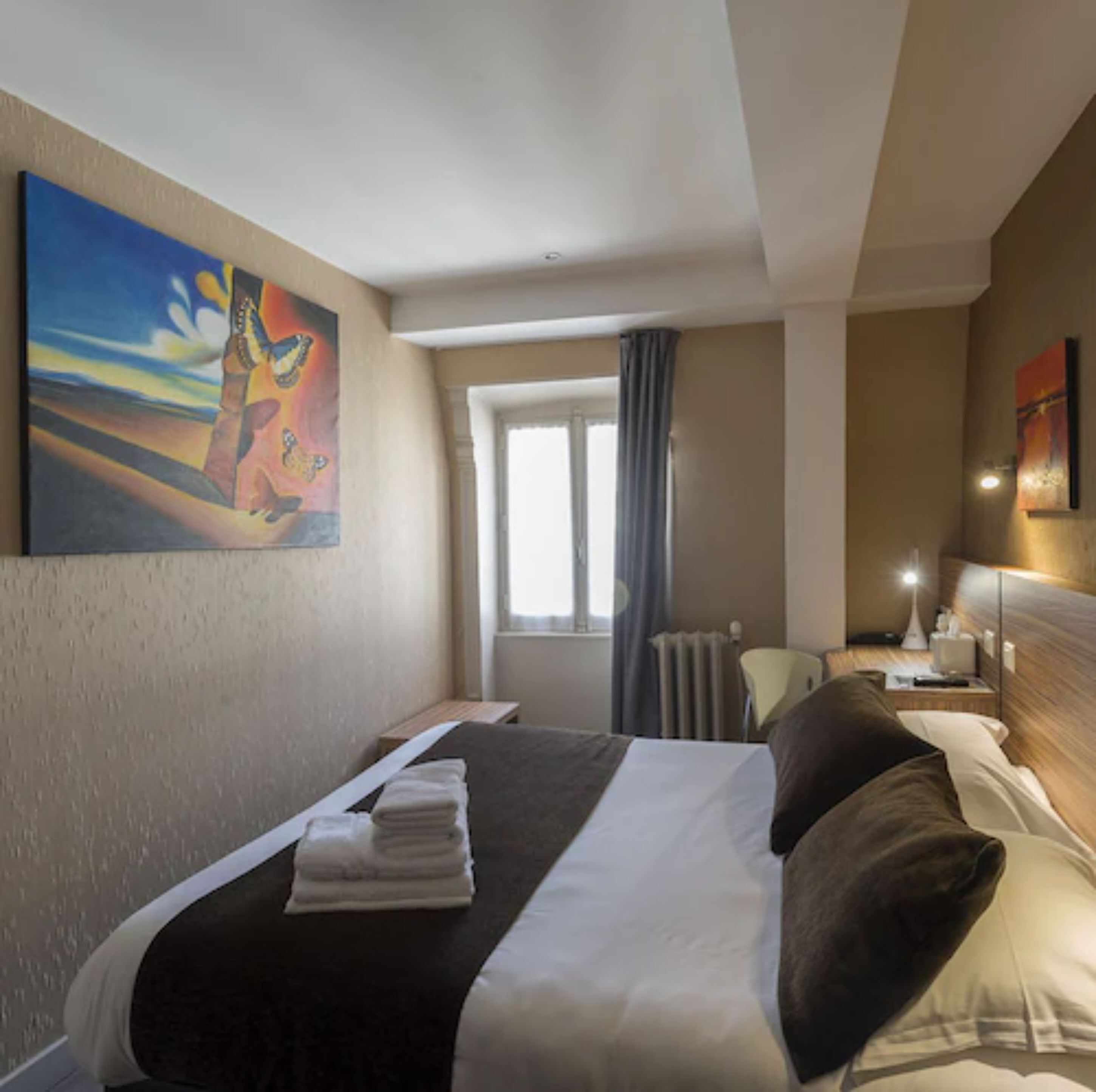 Une chambre d’hôtel avec un lit, une fenêtre, un radiateur, un bureau et un tableau au mur.