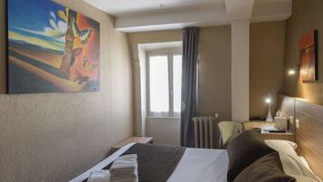 Une chambre d’hôtel avec un lit, une fenêtre, un radiateur, un bureau et un tableau au mur.