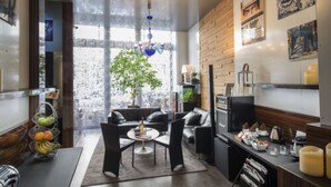 A modern living room with a sofa, coffee table, and a dining area with a table and chairs. There is a large window with a view of trees and a chandelier hanging from the ceiling. - Hôtel de Paris (Lyon)