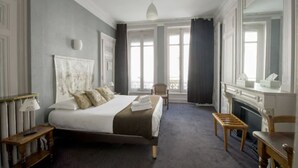 A bedroom with a large bed, a wooden chair, a small table, a fireplace, and a window with curtains. - Hôtel de Paris (Lyon)