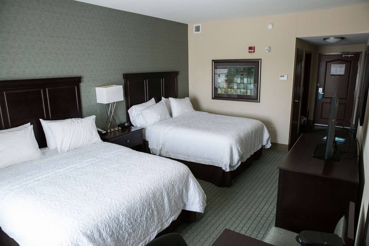 A hotel room with two beds, a desk, a chair, a lamp, and a window with a view.