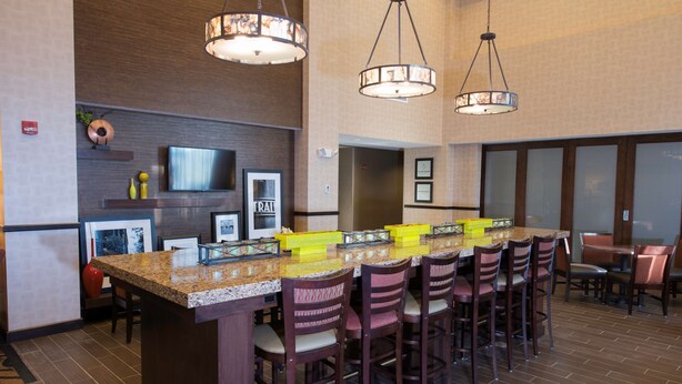 A bar area with a long counter, multiple bar stools, and hanging pendant lights. - Hampton Inn & Suites Bismarck Northwest (Bismarck)