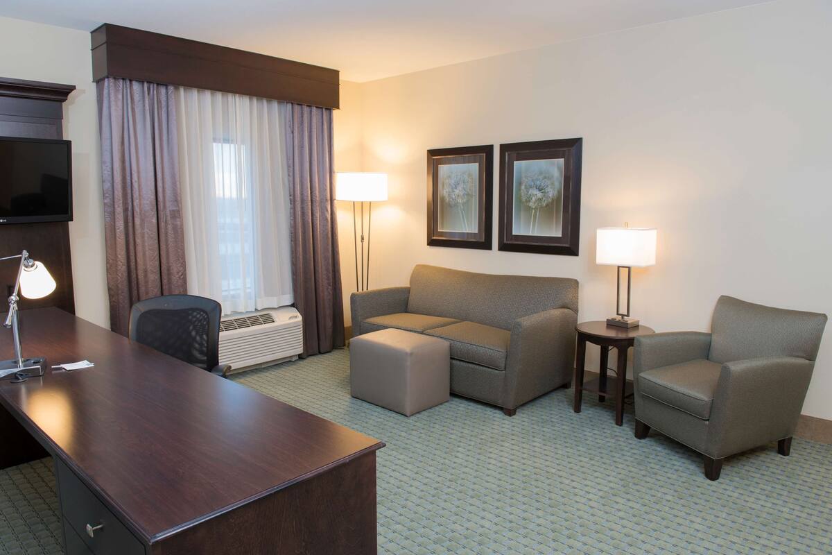 A hotel room with a desk, a sofa, an ottoman, a chair, two lamps, a flat-screen TV, and a window with curtains.