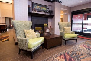 A lobby with two patterned armchairs, a wooden coffee table, a fireplace, and a large window.
