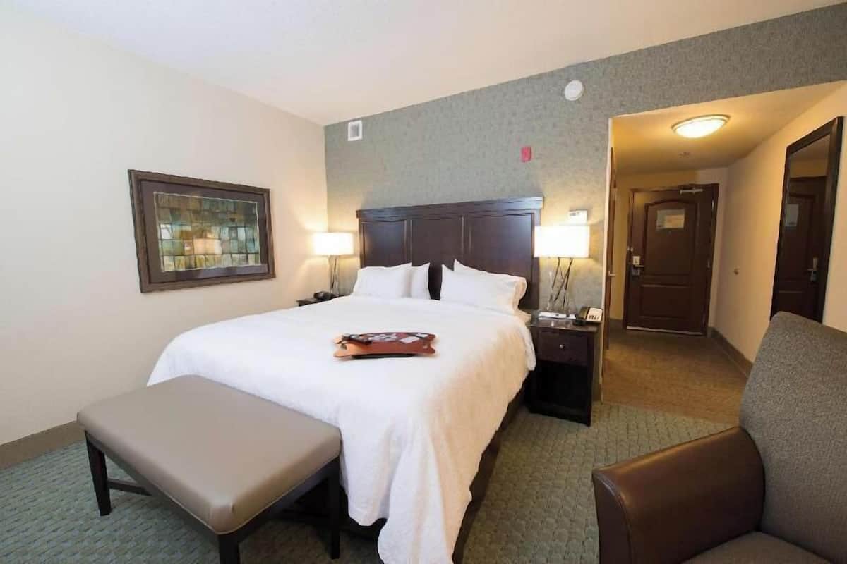 A hotel room with a bed, bedside tables, a bench, a framed picture, and a door leading to another room.
