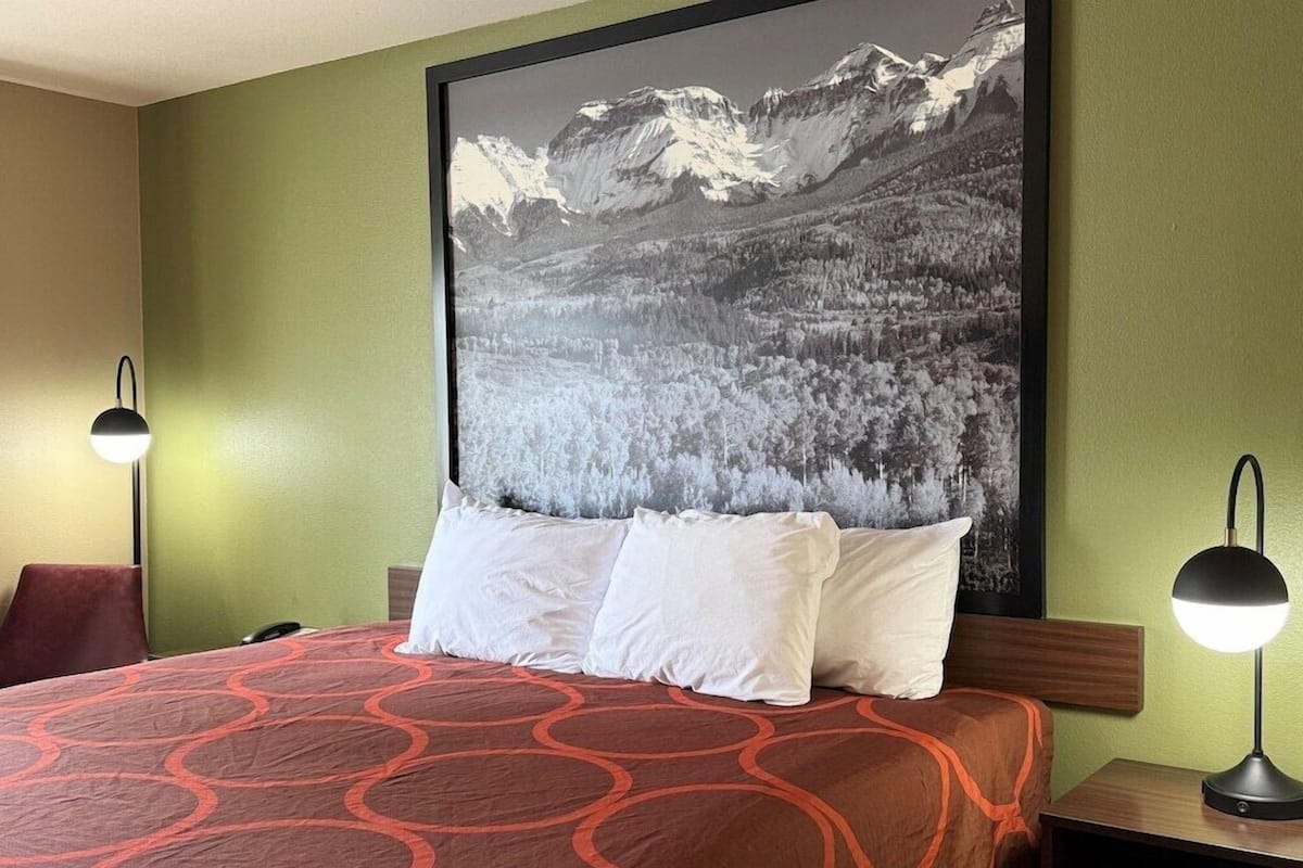 Une chambre d’hôtel avec un grand lit, deux lampes de chevet, un tableau représentant un paysage de montagne accroché au mur et un mur de couleur verte.