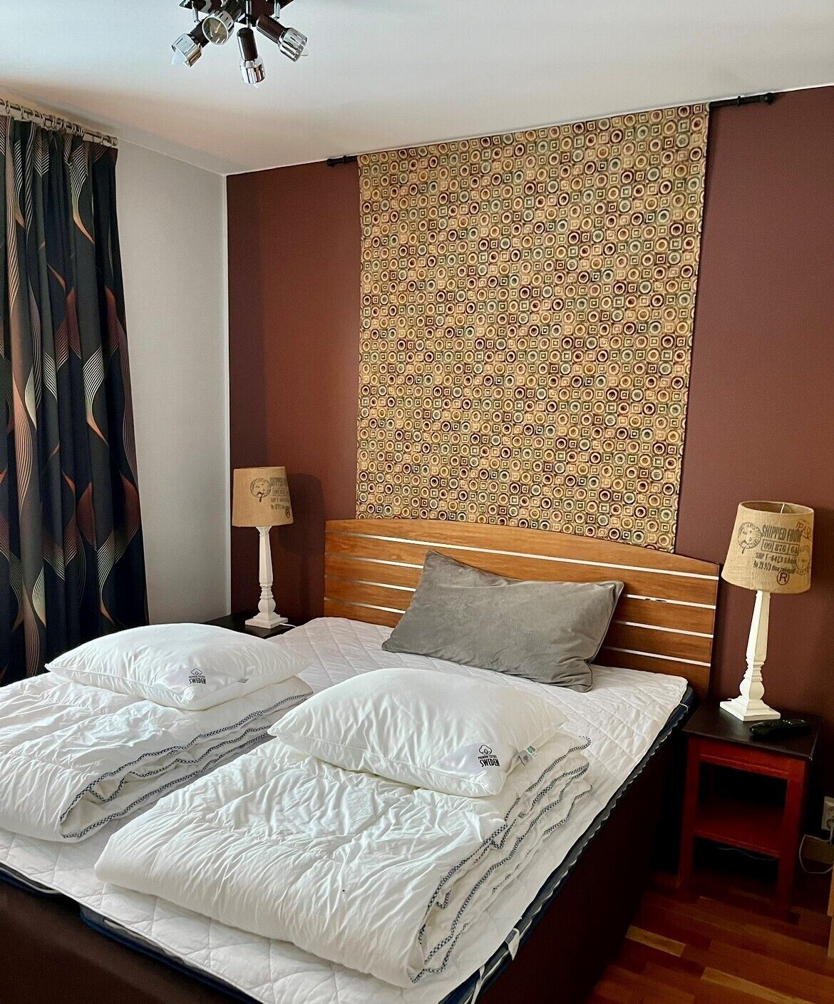 A double bed with white bedding and two pillows, a wooden headboard, a patterned wall, a small bedside table with a lamp, and a curtain with a leaf pattern.