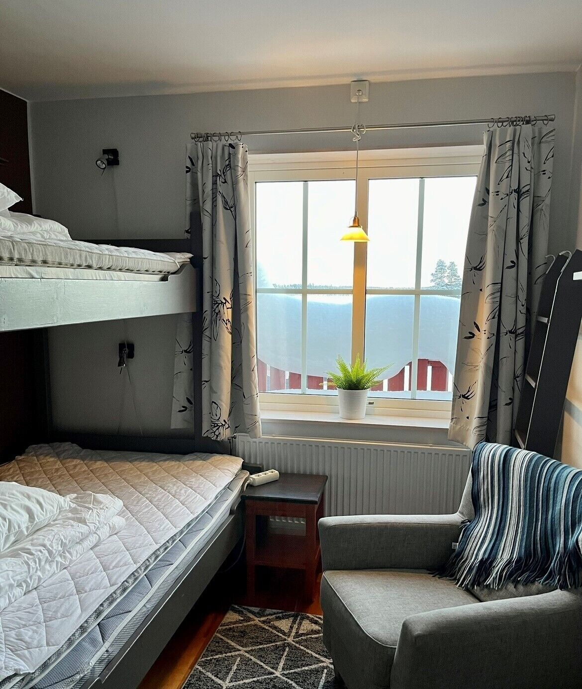 A dorm-style room with bunk beds, a small table, a chair, and a window with a view of a red building and greenery.