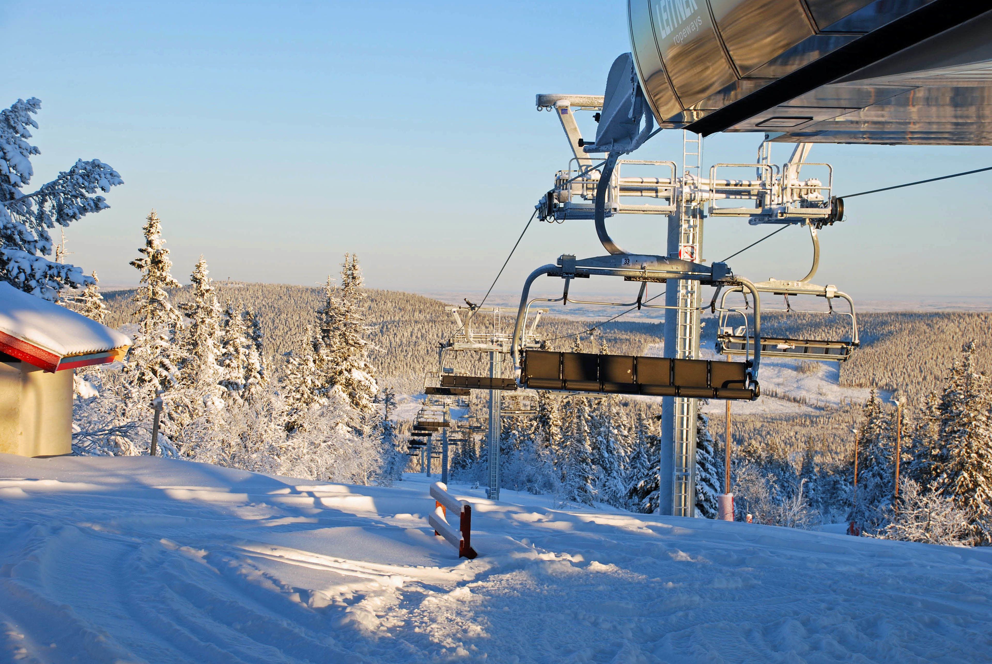 Eine Seilbahn mit Sesseln und Stahlseilen, umgeben von schneebedeckten Bäumen und blauem Himmel.