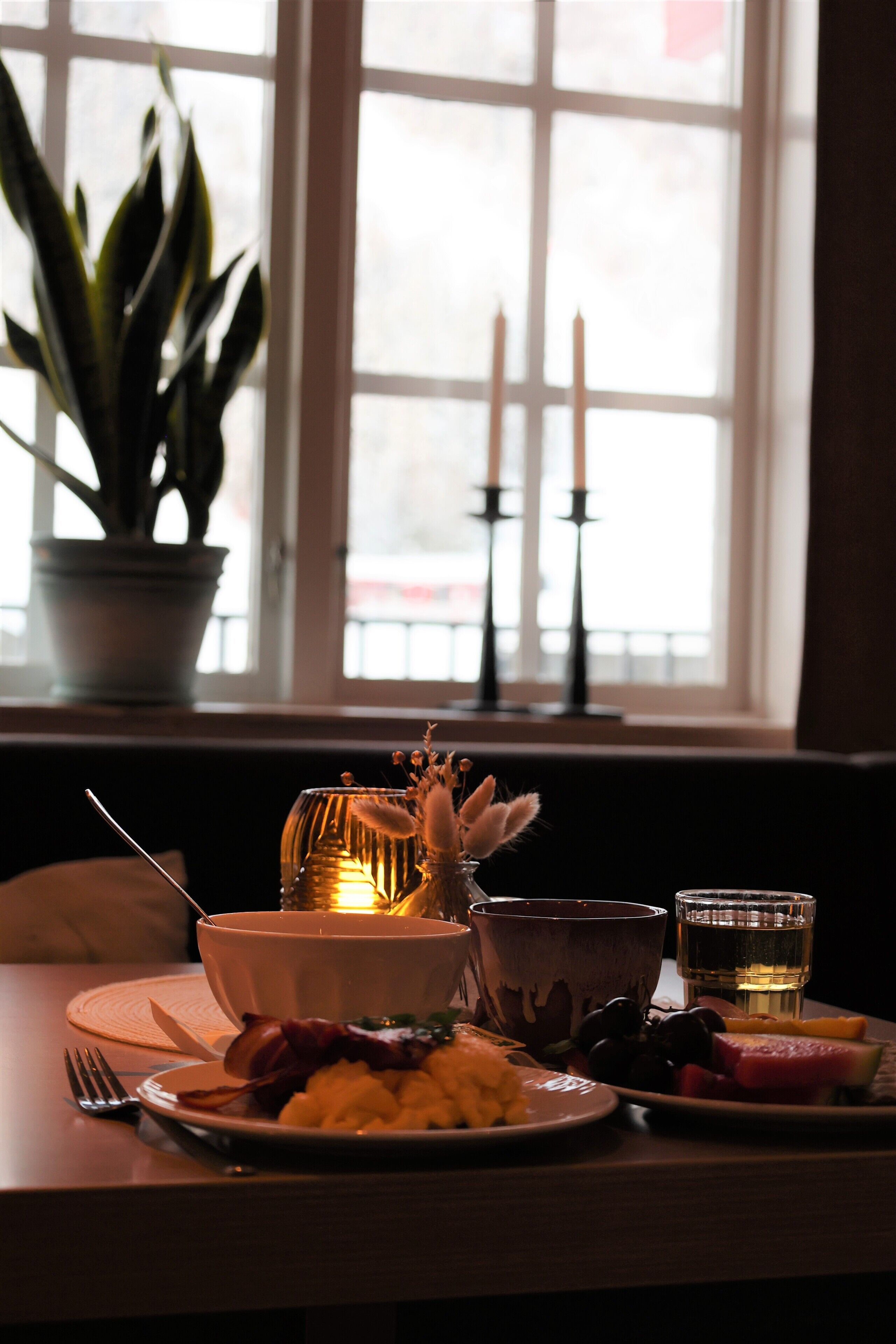 Una mesa de comedor con platos de comida, una vela y una planta en maceta junto a una ventana.