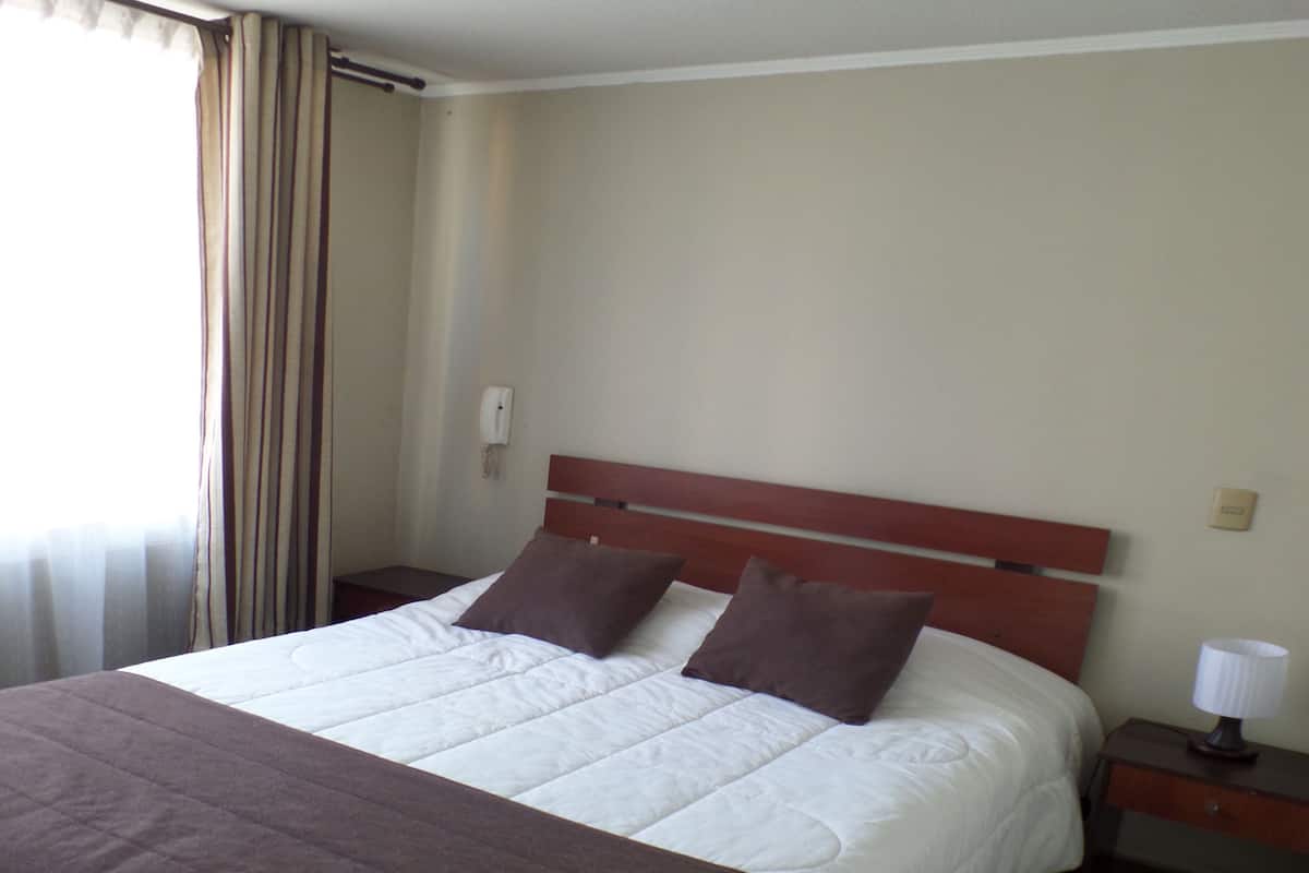 Una habitación de hotel con una cama grande, una mesita de noche, una lámpara y una ventana con cortinas.