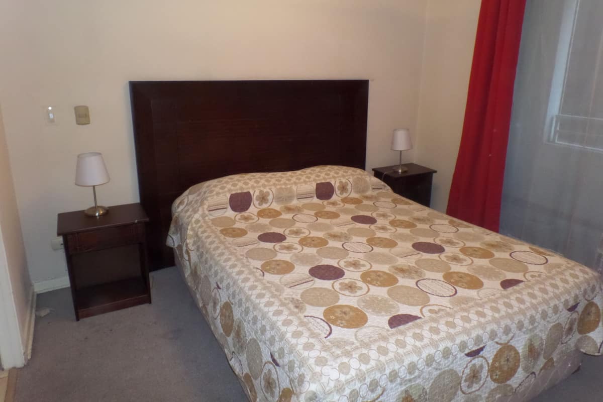 Una cama bien hecha con una colcha estampada, dos mesitas de noche con lámparas y una cortina roja en la ventana.