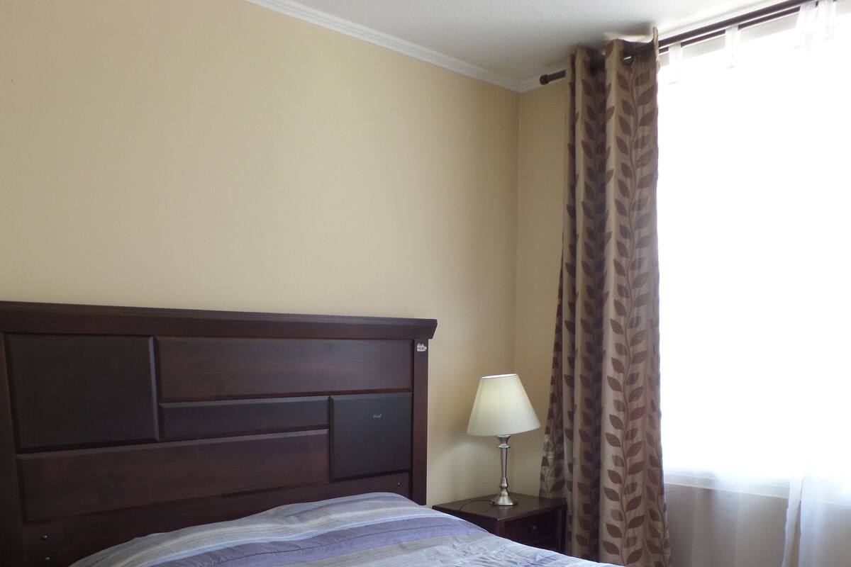 Una habitación de hotel con una cama grande, una mesita de noche, una lámpara y un ventanal con cortinas.