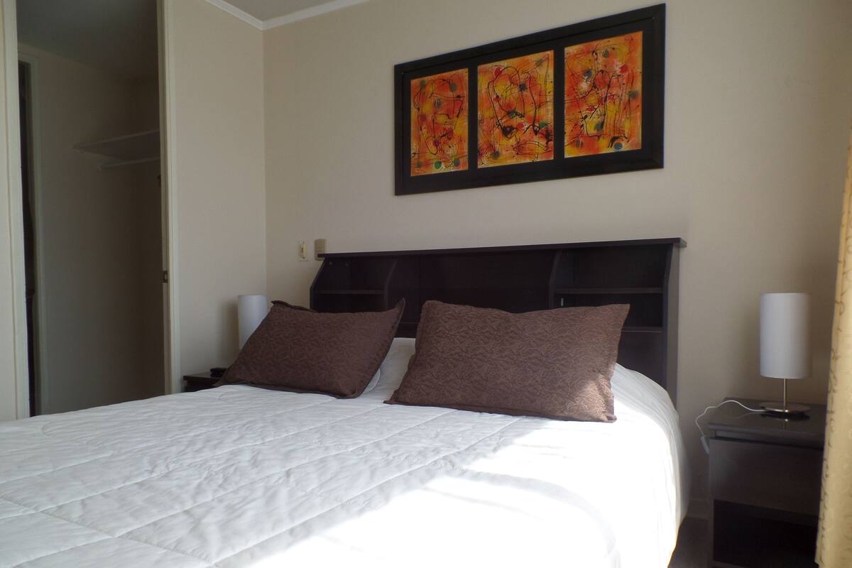 Una cama bien hecha con edredón blanco y dos almohadas marrones, una mesita de noche con lámpara y un cuadro sobre la cama.