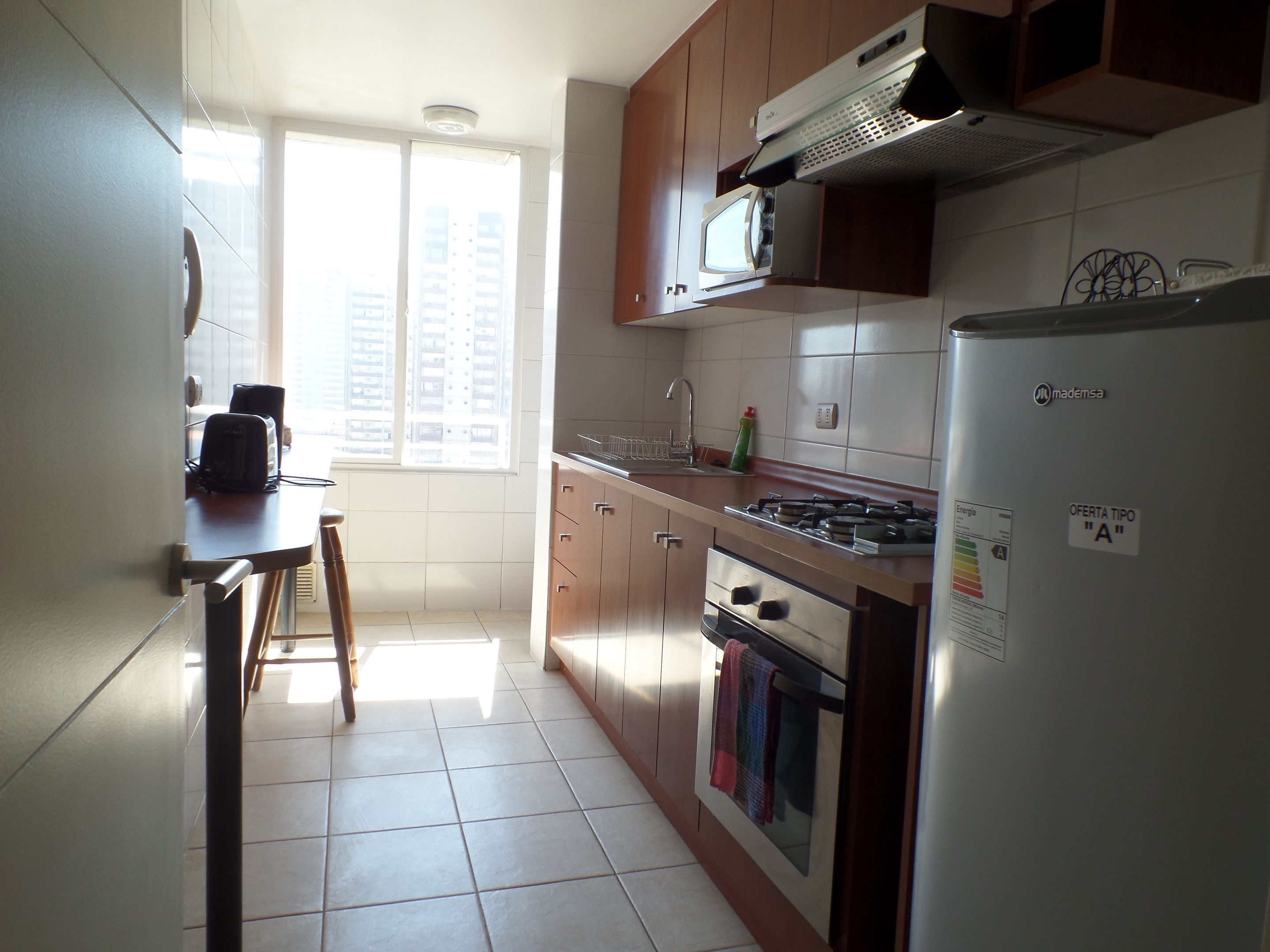 Een keuken met een fornuis, magnetron en koelkast. Er is een raam dat natuurlijk licht en uitzicht op gebouwen biedt.