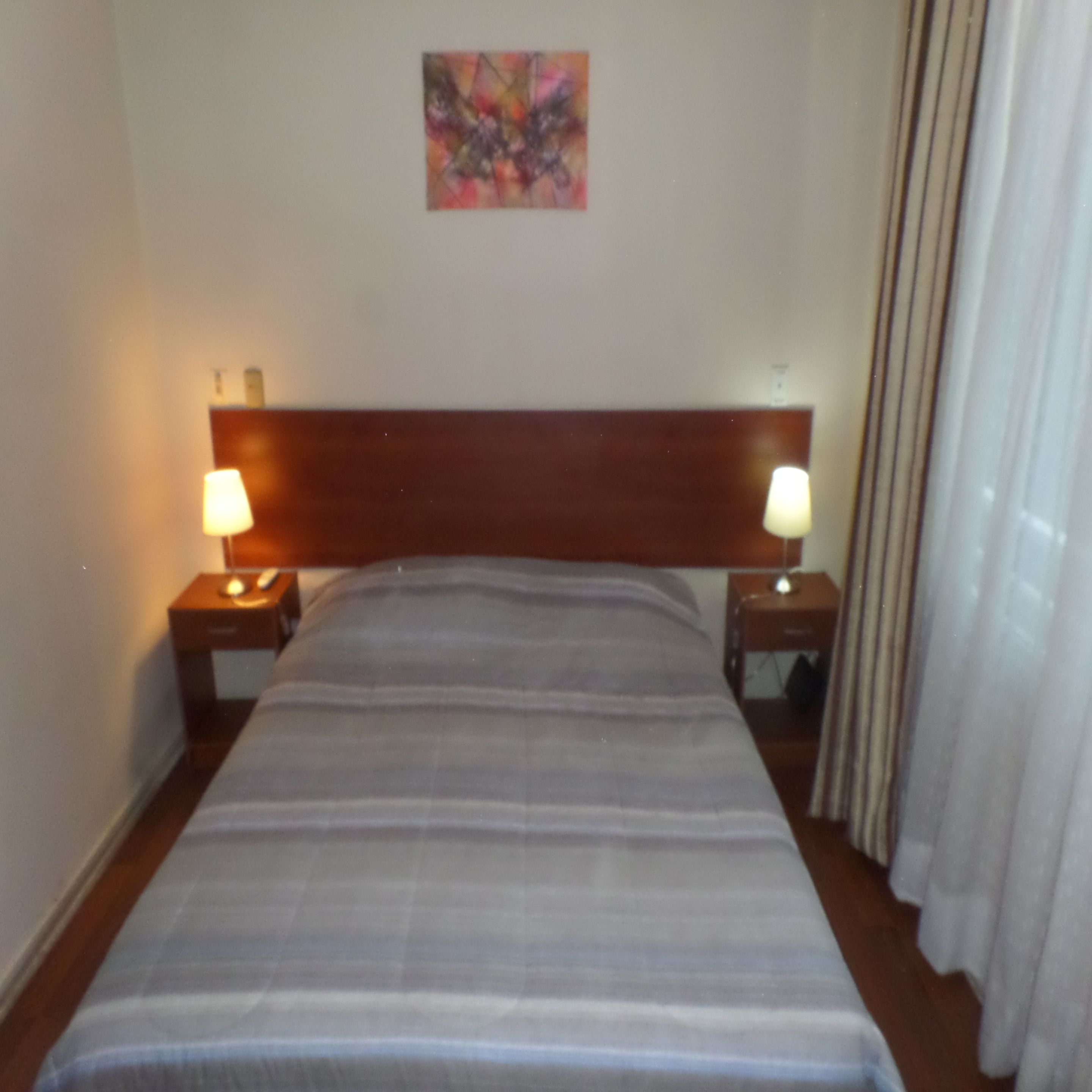 A hotel room with a bed, two bedside lamps, a nightstand, a painting on the wall, and a window with curtains.