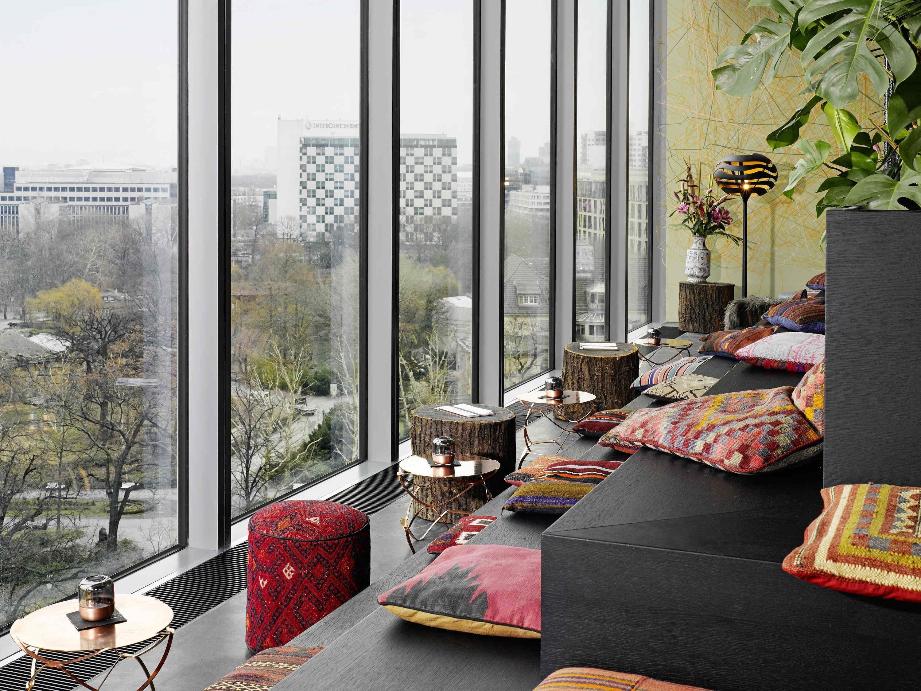 A modern living room with large windows, a black sofa, colorful cushions, and a view of a cityscape.