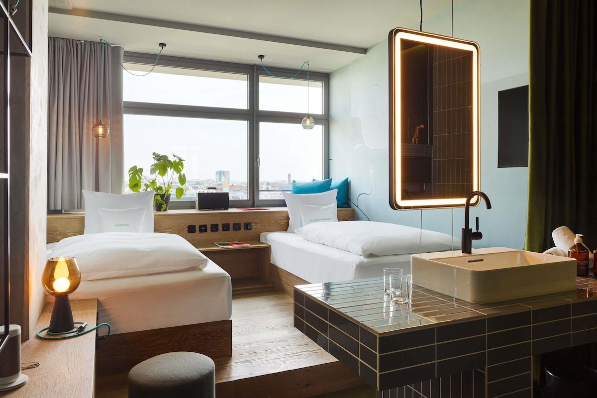 Una habitación de hotel moderna con dos camas, un ventanal grande, una mesita de noche con lámpara, un espejo y un baño con lavamanos y artículos de aseo.