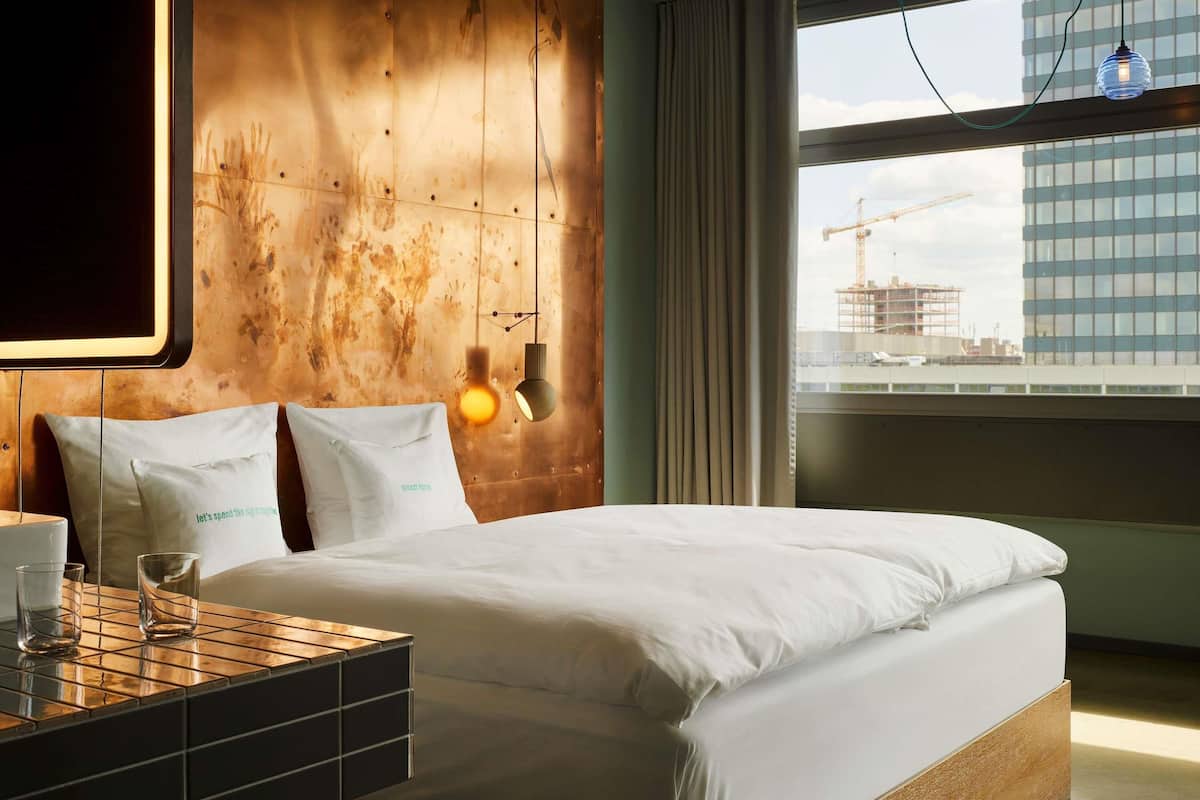 A modern hotel room with a large bed, a wooden headboard, a bedside table with glasses, and a view of a cityscape through a large window.