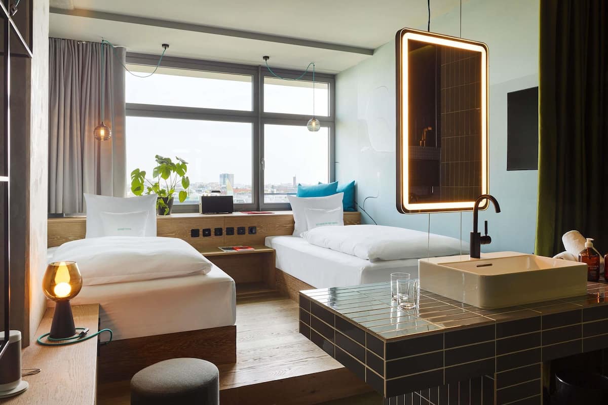 A modern hotel room with two beds, a large window, a bedside table with a lamp, a mirror, and a bathroom with a sink and toiletries.