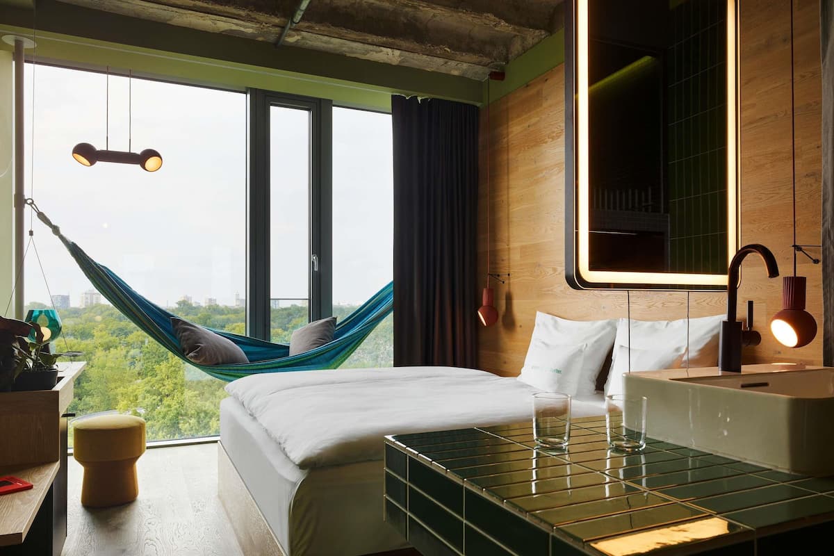 A modern hotel room with a large bed, a view of greenery, a hammock, a bedside table with a lamp, and a bathroom with a mirror and sink.