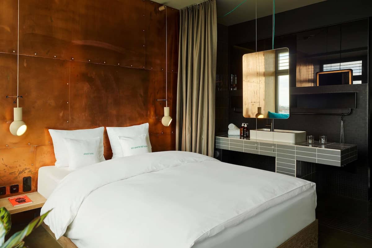 A hotel room with a large bed, a bedside table with a lamp, a mirror, and a modern bathroom with a sink and mirror.