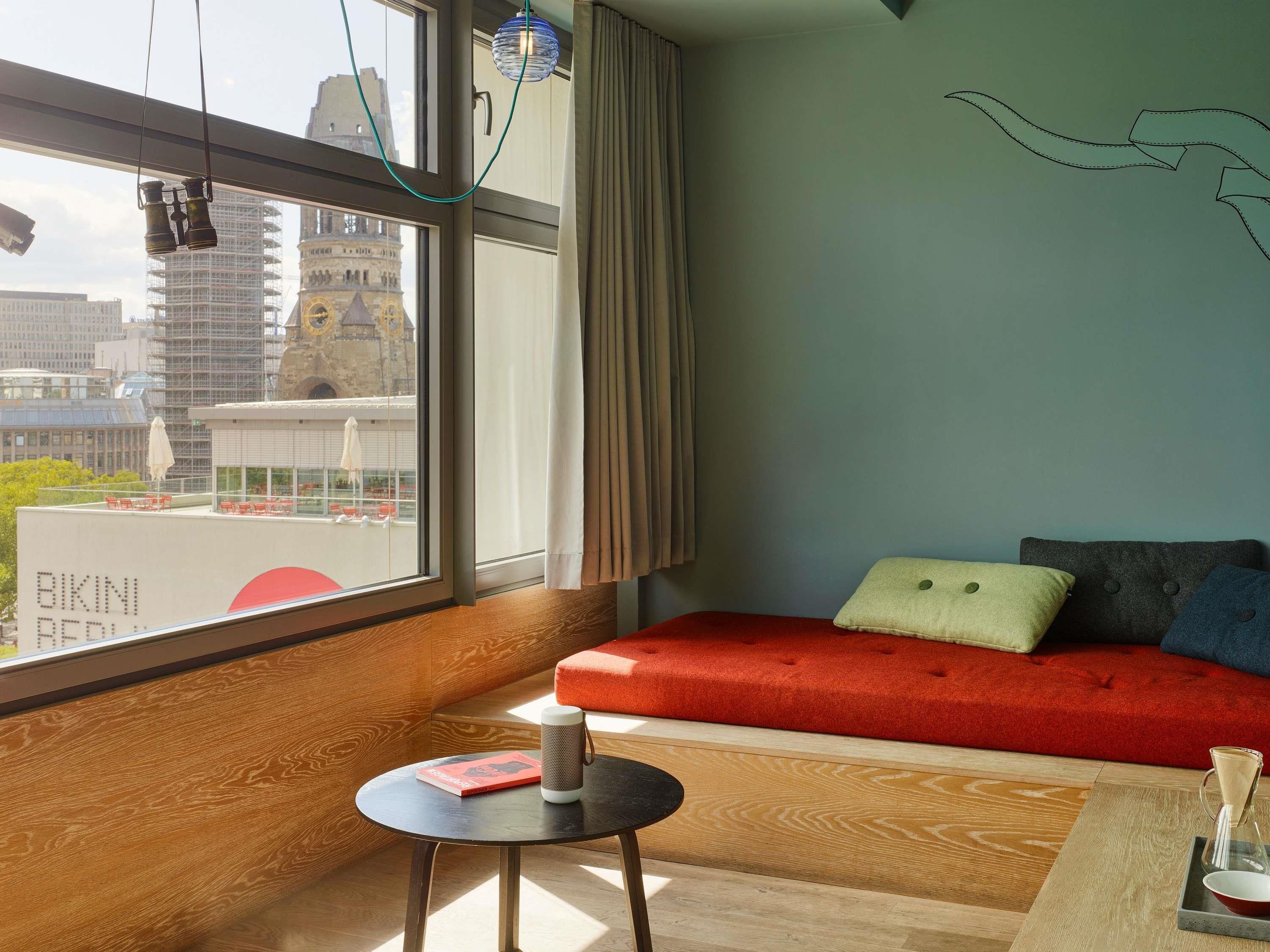 A modern room with a large window, a red sofa, a small table with a book and a speaker, and a view of a cityscape.