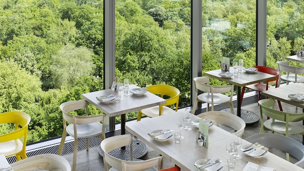 A restaurant with tables set for dining, featuring a view of a lush green landscape through large windows. - 25hours Hotel Bikini Berlin (Berlin)