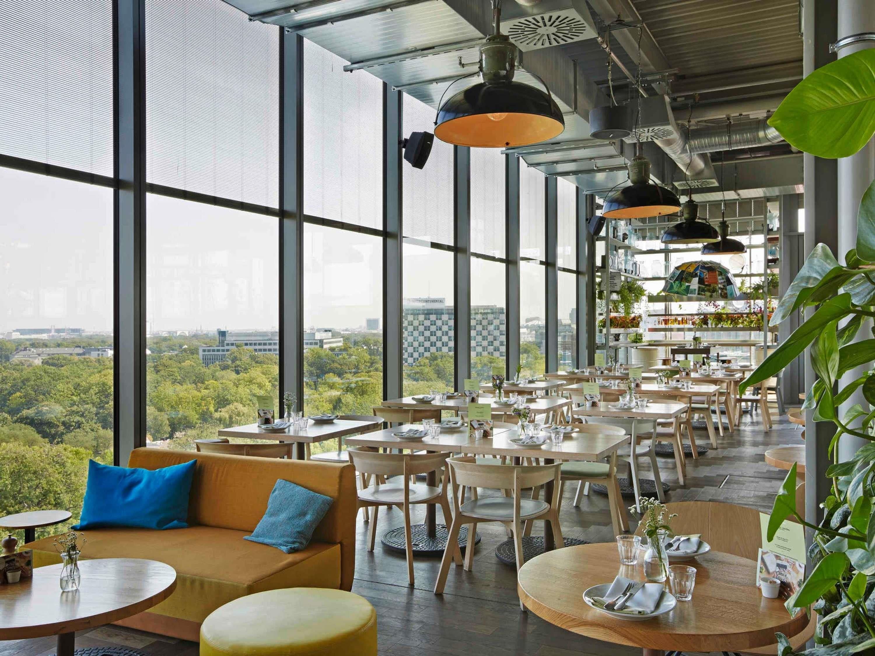 A restaurant with large windows offering a city view, wooden furniture, and hanging pendant lights.