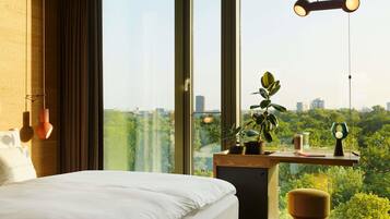 Une chambre moderne avec un grand lit, un bureau agrémenté d’une plante en pot et une vue sur le paysage urbain, visible à travers une grande fenêtre.