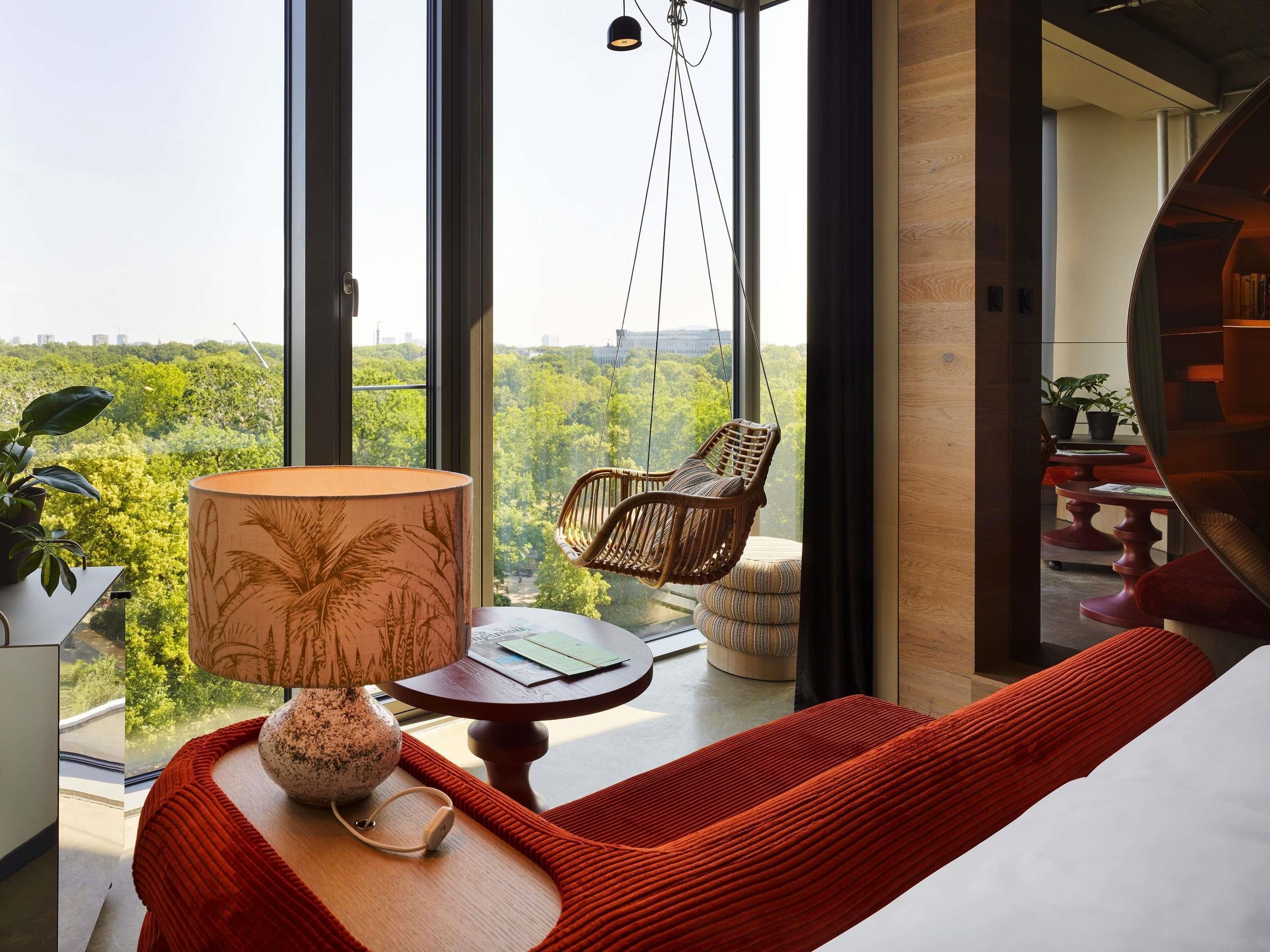 Una habitación con un ventanal grande, una silla colgante, una mesa con lámpara y un sofá rojo.