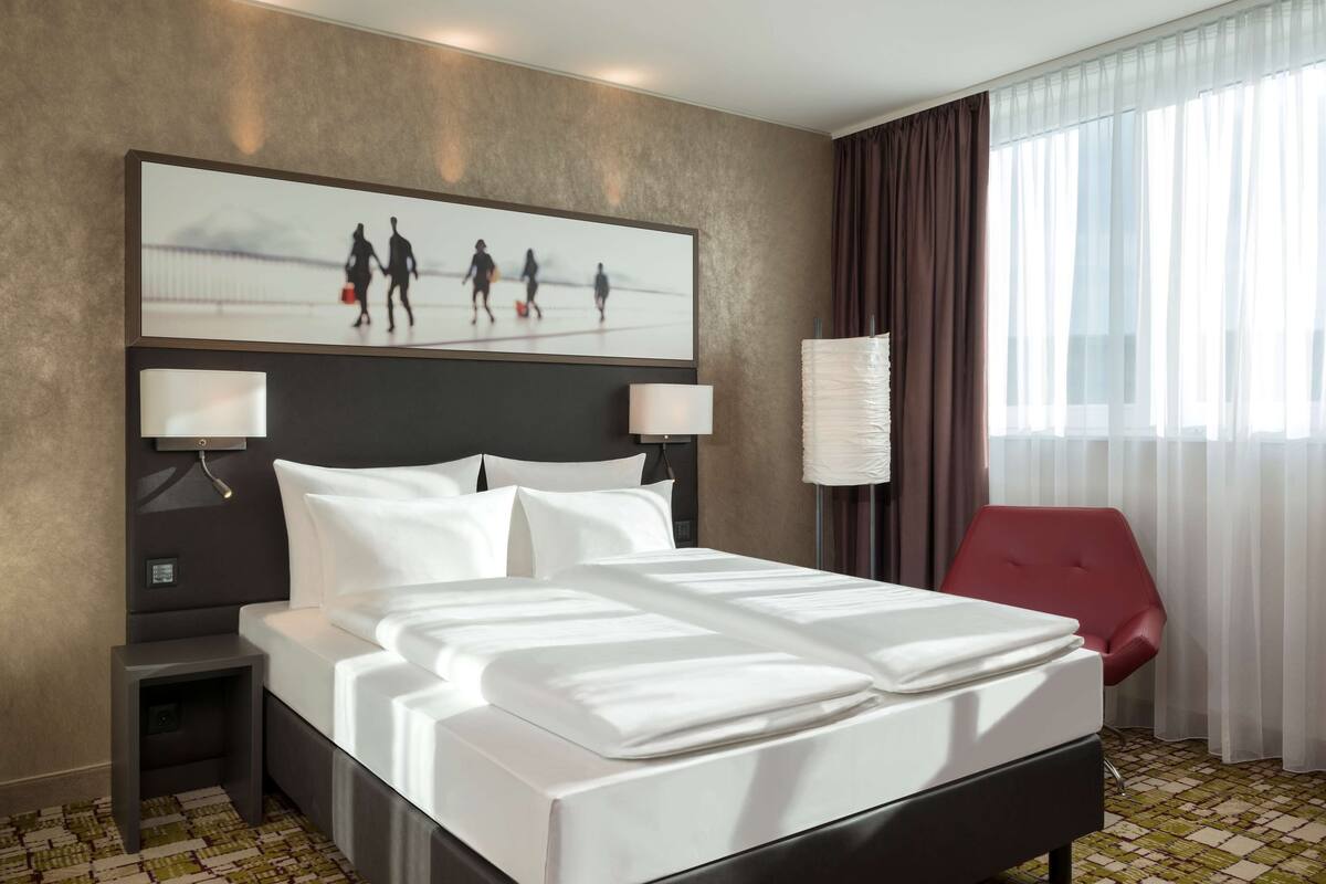 A hotel room with a large bed, two bedside tables with lamps, a red chair, a large window with curtains, and a painting on the wall.